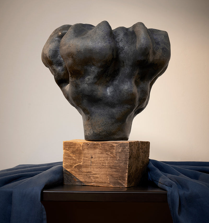 Earthy amorphous vase in indigo and copper glaze on top on a wooden block arranged on a tabletop with a dusty washed navy tablecloth. 